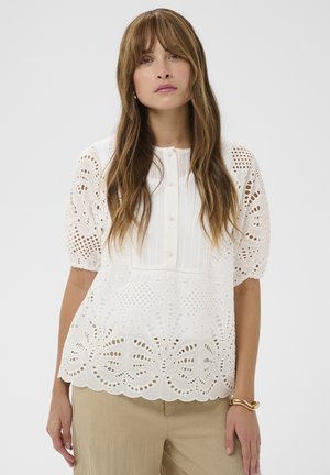 Kobieta o długich brązowych włosach ubrana w białą bluzkę z krótkim rękawem z ażurowej koronki oraz beżowe spodnie, stojąca na jednolitym tle.