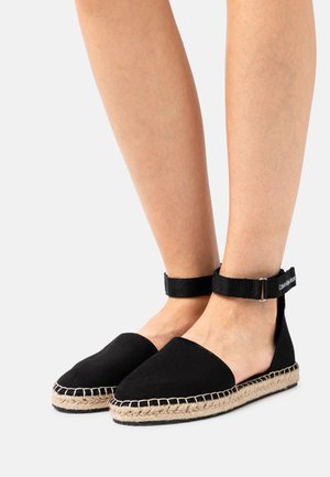 Espadrilles noires en toile avec une semelle en jute, dotées d'une sangle en tissu à la cheville et d'un design ouvert à l'arrière. Détails de couture le long du bord.
