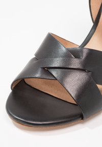 Sandale en cuir noir avec des brides croisées et une finition lisse. Elle présente un bout arrondi et une semelle intérieure texturée pour un confort accru.