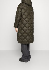 Olivově zelená, péřová bunda s kapucí a prošívaným diamantovým vzorem, dlouhými rukávy, sahající po lýtka, kombinovaná se širokými šedými džíny a černými botami.