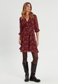 Robe florale bourgogne à manches longues, taille cintrée et ourlet froncé. Associée à des bottes en cuir noir jusqu'aux genoux et des collants foncés.