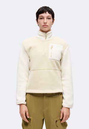 Person mit kurzen dunklen Haaren, die einen cremefarbenen Fleece-Pullover mit Reißverschluss-Brusttasche und olivgrüne Hosen mit Reißverschlusstaschen trägt.