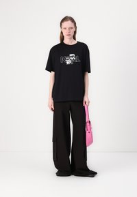 Black oversized t-shirt with a cartoon graphic and text "KARL"; paired with loose black pants. Holding a small pink handbag.