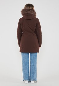 Parka isolato marrone con rifinitura in pelliccia sintetica sul cappuccio, texture liscia e design leggermente svasato, abbinato a jeans blu a gamba larga e sneaker bianche.