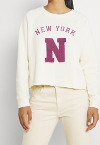 Donna che indossa una felpa panna con la scritta "New York College" e una grande "N" viola, abbinata a pantaloni a righe beige chiaro, vista dal collo ai fianchi.