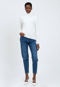 Maglione bianco a coste con collo alto abbinato a jeans blue a gamba dritta. Il modello indossa tacchi bianchi a punta con cinturini alla caviglia. Look semplice e moderno.