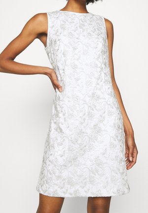 Robe blanche sans manches avec broderie florale argentée portée par une personne la main sur la hanche, debout devant un fond blanc.