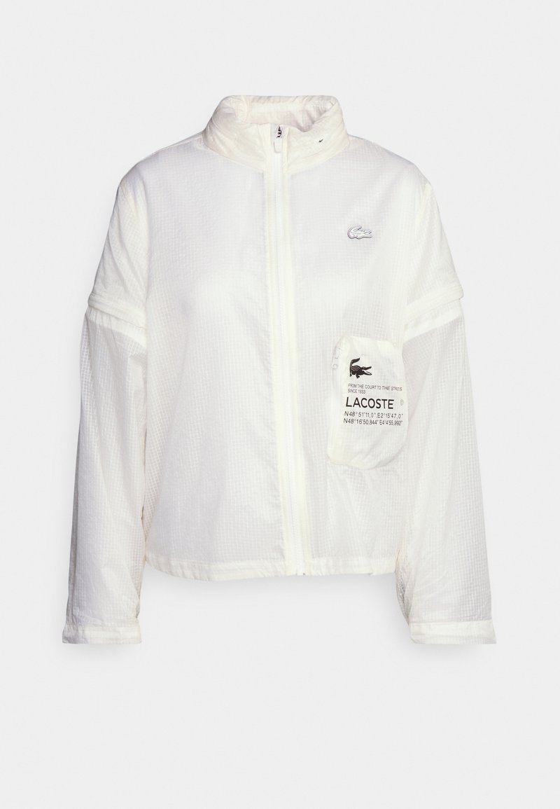 Coupe-vent léger de couleur blanche avec une surface texturée, fermeture éclair intégrale, poches latérales et logo Lacoste sur la poitrine. Possède des manches courtes.