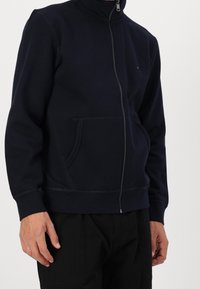 Sudadera con cremallera de color azul marino oscuro, fabricada en tejido de mezcla de algodón. Presenta un collar alto, dos bolsillos frontales y puños ribeteados en el bajo.