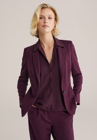 Lila Blazer mit Reverskragen, einzelnem Knopfverschluss, kombiniert mit einem passenden lila Hemd mit durchgehendem Knopfverschluss und strukturiertem Stoff.