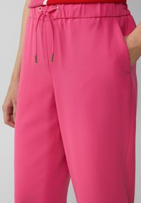 Roze joggerbroek gemaakt van een gladde stof met een elastische tailleband, een trekkoord met metalen tips en zijzakken.
