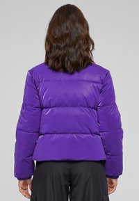 Fioletowa puchowa kurtka o pikowanym wzorze, z poziomymi sekcjami, dopasowaną talią i elastycznymi mankietami. Gładka, błyszcząca tekstura.