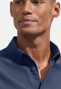 Tumši zilas krāsas teksturēta krekla ar pogām uz apkakles, kas satur divas pogas un gludu virsmu. Tuvplāns uz apkakles un kakla daļu.