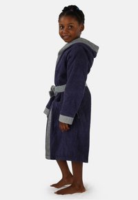 Lila Frottee-Bademantel mit einer grauen Kapuze und Säumen, ausgestattet mit einem Gürtel und Taschen an der Seite, getragen von einem barfuß stehenden Kind.