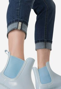 Vaaleansiniset kumiset nilkkasaappaat, joissa on elastiset sivupaneelit, yhdistettynä rullattuihin tummiin farkkuihin, joissa on kontrastivärinen sininen helma.