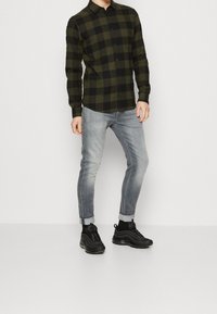 Camisa de cuadros en negro y verde, jeans slim-fit de color azul claro y zapatillas negras. La camisa tiene cuello y mangas largas.