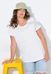 Camiseta blanca de manga corta con bordados en forma de estrella en el hombro. El modelo lleva pantalones cortos de mezclilla azul claro y un sombrero tipo cubo verde claro.