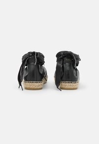 Espadrilles de couro preto com bico arredondado, apresentando uma sola de juta trançada e grandes fitas de cetim atadas na parte de trás. Acabamento brilhante.