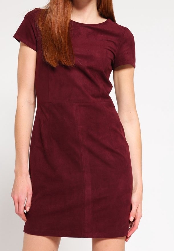 Femme aux longs cheveux roux portant une robe en daim bordeaux à manches courtes, debout devant un fond clair uni.