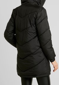 Svart puffjacka med huva, med ett kviltat chevronmönster, elastiska ärmslut och en något böjd nederkant.