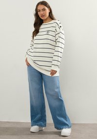 Vit randig sweatshirt med marinblå ränder, knappdetaljer på axeln, i kombination med vida ljusblå jeans och vita sneakers.