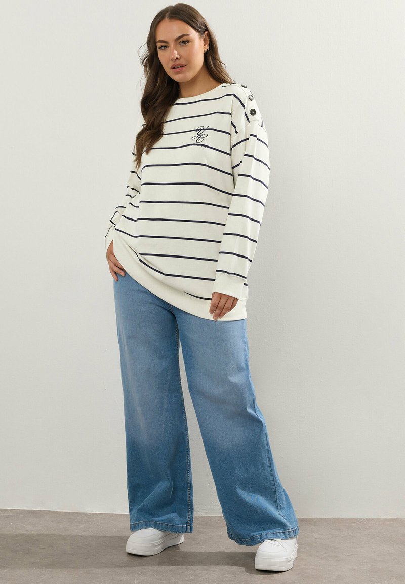 Vit randig sweatshirt med marinblå ränder, knappdetaljer på axeln, i kombination med vida ljusblå jeans och vita sneakers.