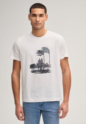 Junger Mann, der ein weißes T-Shirt mit schwarzem Palmen- und Surfer-Silhouetten-Motiv trägt, kombiniert mit hellblauen Jeans, stehend vor einfachem Hintergrund.