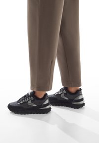 Le sneakers nere e grigie presentano una tomaia testurizzata con accenti riflettenti e un colletto soffice, poggiati su una suola nera robusta.