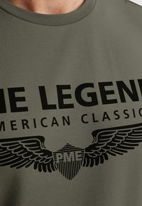 Olivově zelené bavlněné tričko s velkým černým textem a grafikou štítu s křídly. Kulatý výstřih a krátké rukávy.