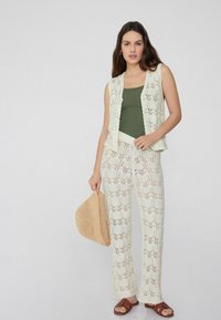 Gehaakt vest en broek in off-white met bladmotieven, gecombineerd met een groene tanktop en bruine sandalen, terwijl een strohoed wordt vastgehouden.