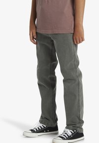 Jeans de denim gris con corte recto y detalles de costura mínimos, combinados con zapatillas bajas negras que presentan cordones y acentos blancos.