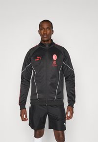 Sort atletisk jakke med røde accenter og hvid piping, som har høj krave, lynlåslukning og logoer på brystet og siden.