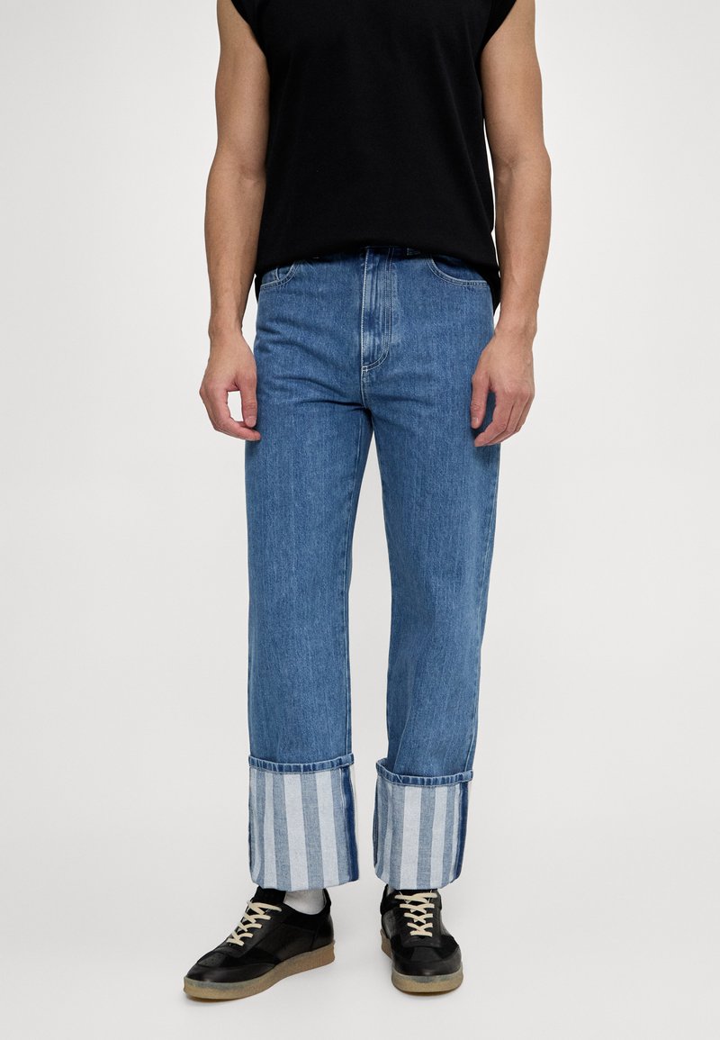 Jeans de denim azul con un corte holgado, que presentan puños rayados en azul claro y blanco. Combinados con zapatillas negras y una camiseta sin mangas negra.
