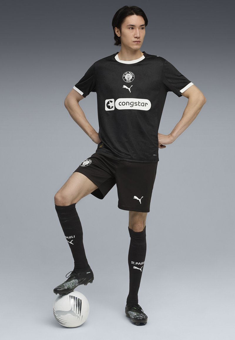 Jauns vīriešu futbolists melnā St. Pauli formā pozē ar vienu kāju uz balta futbola bumbas pret pelēku fonu.