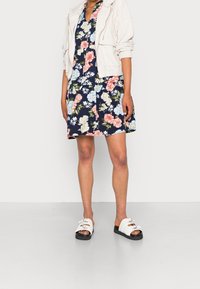 Robe florale avec un fond marine, ornée de fleurs roses et bleues. Accompagnée d'une veste texturée gris clair et de sandales à plateforme blanches.