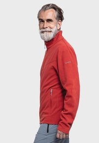 Rote Fleecejacke mit hohem Kragen, halbem Reißverschluss vorne, Seitentaschen und gesticktem Logo am Ärmel. Glatte Textur und warmes Erscheinungsbild.