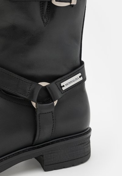 Botín negro de cuero hasta el tobillo con una hebilla de tono plateado y un acento circular, con una suela texturizada y detalles de costura. Etiqueta de la marca visible.