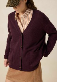 Cardigan V à col en laine de couleur violet foncé, avec des boutons et deux poches avant. Associé à une chemise à rayures à boutons et à une jupe en velours côtelé.