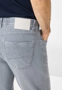 Hellgraue Jeanshose mit glatter Textur, ausgestattet mit einem weißen Markenlabel am Bund und einer Hintertasche mit minimaler Naht.