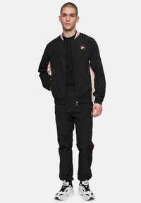 Svart bomberjacka med röda och beige sidestreck, med rund krage och dragkedja. Tillsammans med svarta joggers och vita sneakers.