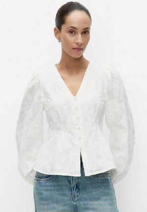 Nainen, jonka hiukset ovat sidottu taakse, pukeutuneena valkoiseen kukkakirjailtuun napitettuun puseroon, jossa on ilmavat pitkät hihat, ja sinisiin farkkuihin.