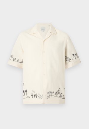 Man with short blonde hair wearing a cream short-sleeve button-up shirt with black tropical prints and light blue wide-leg jeans.