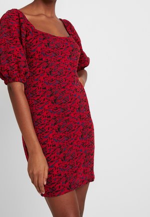 Femme portant une mini robe rouge texturée avec des manches courtes bouffantes et un décolleté carré, se tenant devant un fond clair uni.