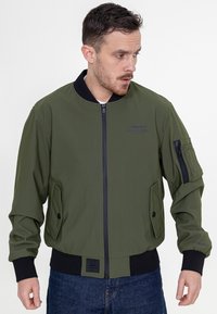 Oliwkowa bomberka z czarnym prążkowanym kołnierzem, zapinana na zamek błyskawiczny, z bocznymi kieszeniami i kieszenią na ramieniu; gładki materiał.