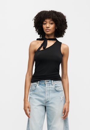 Νεαρή γυναίκα με κατσαρά μαλλιά που φοράει μαύρο ασύμμετρο αμάνικο τοπ με δετό λαιμό και ανοιχτό μπλε τζιν ψηλόμεσα σκισμένα παντελόνια.