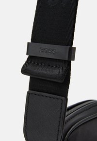 Sangle de sacoche en nylon noir avec un ajusteur en plastique marqué "BOSS" et un accent en cuir renforçant le point d'attache.