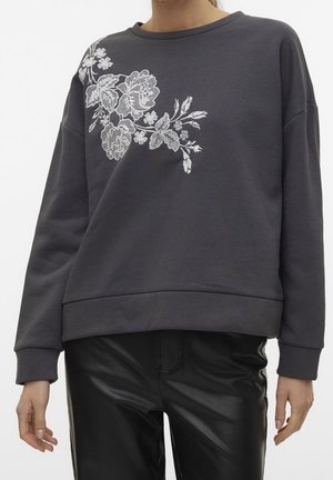Sweat-shirt gris foncé avec broderie florale blanche sur la partie supérieure gauche de la poitrine, associé à un pantalon en cuir noir.