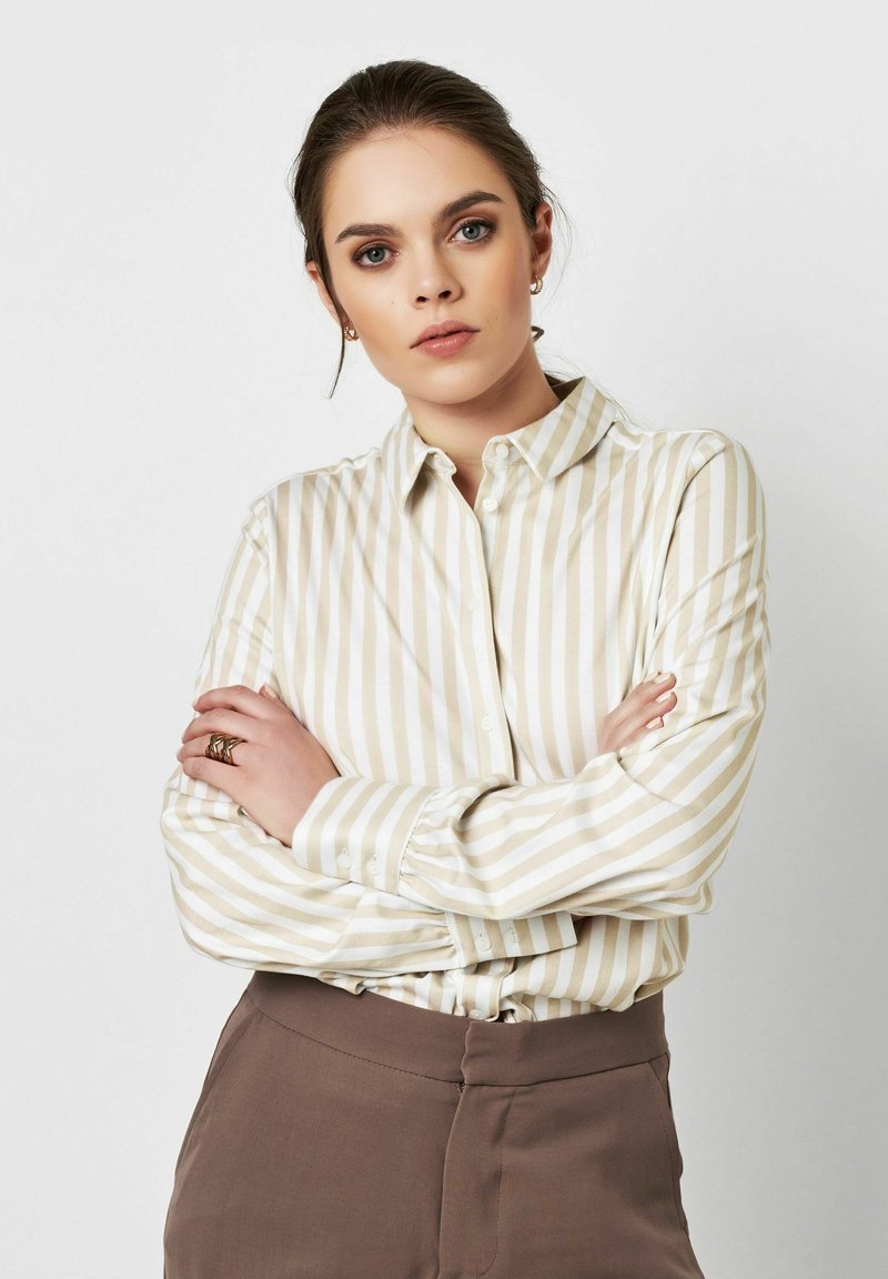 Light beige button-up shirt with white vertical stripes; features long sleeves, a collar, and a smooth texture. Paired with brown trousers.