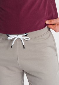 Pantalones de chándal color beige claro con un cordón blanco, que presentan terminales negros, combinados con una camiseta color burdeos. Primer plano de la cintura y la prenda superior.