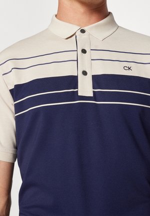 Mann trägt ein beige und marineblau gestreiftes Poloshirt mit dreiknöpfiger Knopfleiste und kleinem "CK"-Logo auf der Brust.